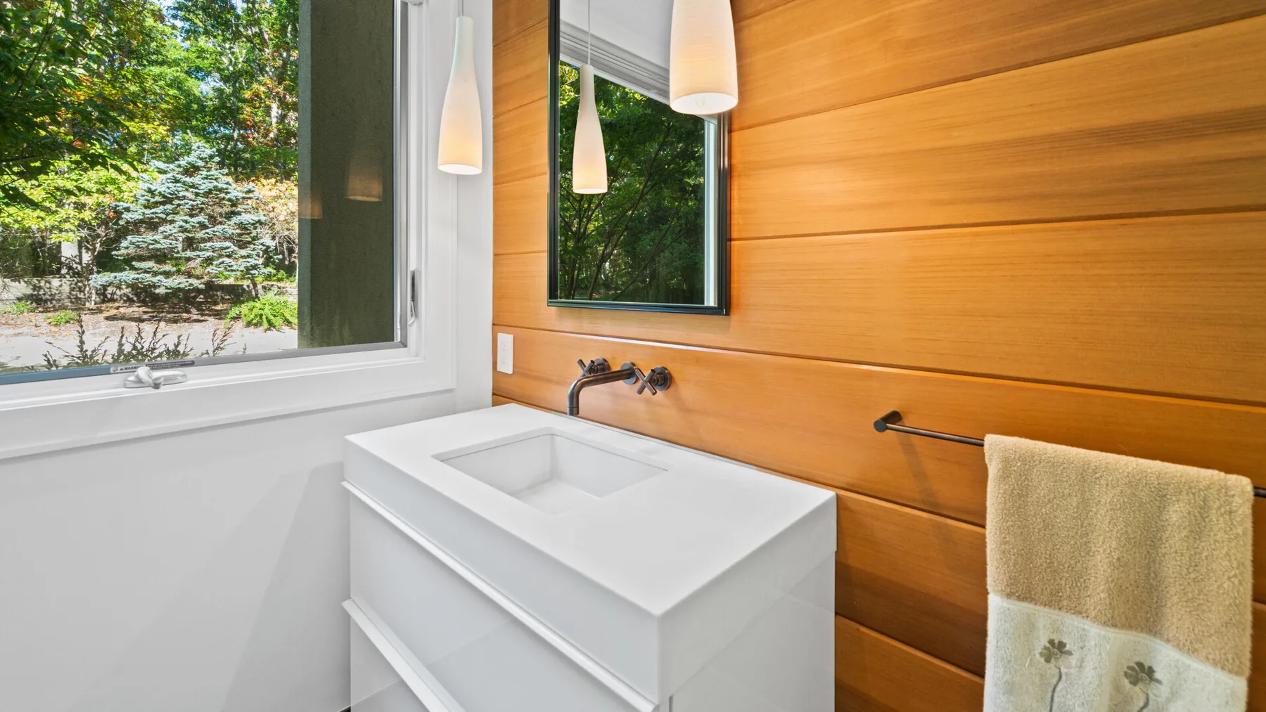 A bathroom vanity sits beside a large window overlooking green trees.