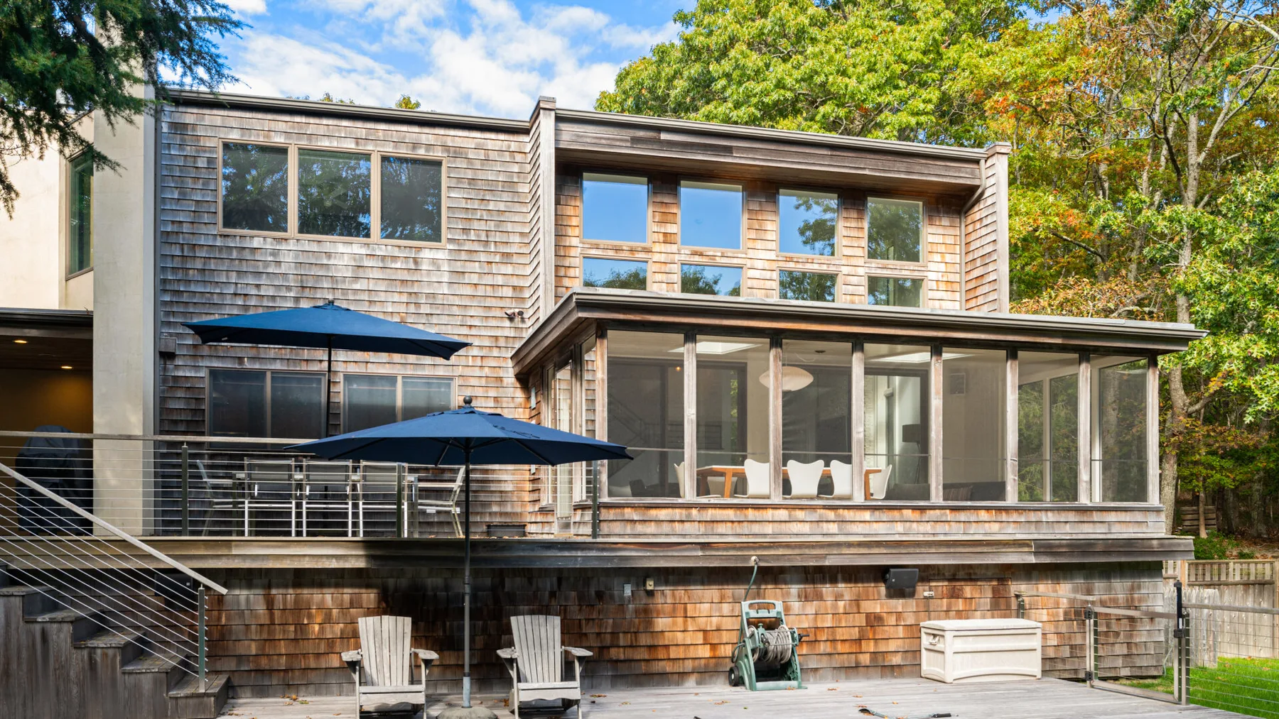 A modern house with a broad deck and outdoor seating, surrounded by trees.