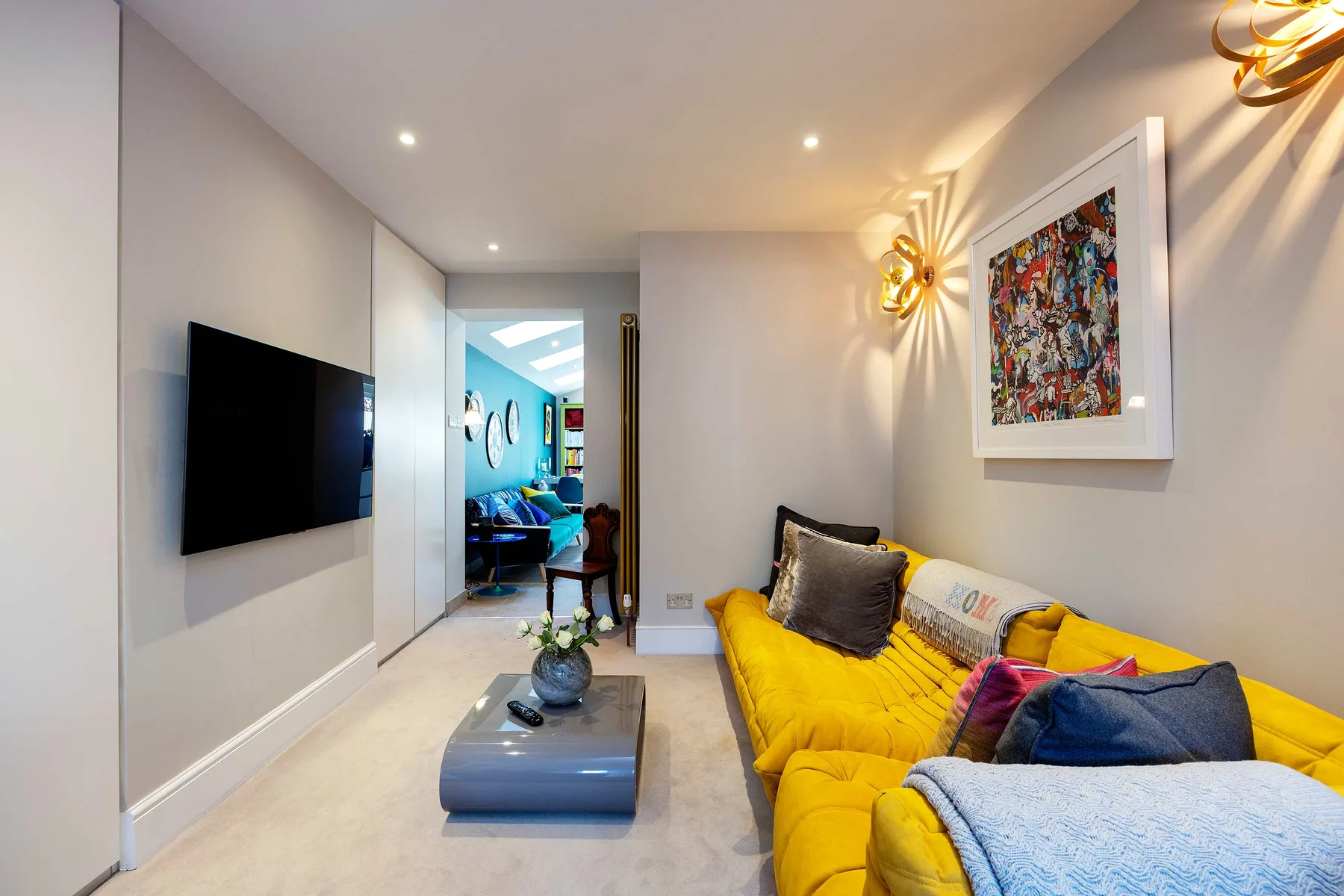 Una sala luminosa con un sofá amarillo, un televisor en la pared y una obra enmarcada.