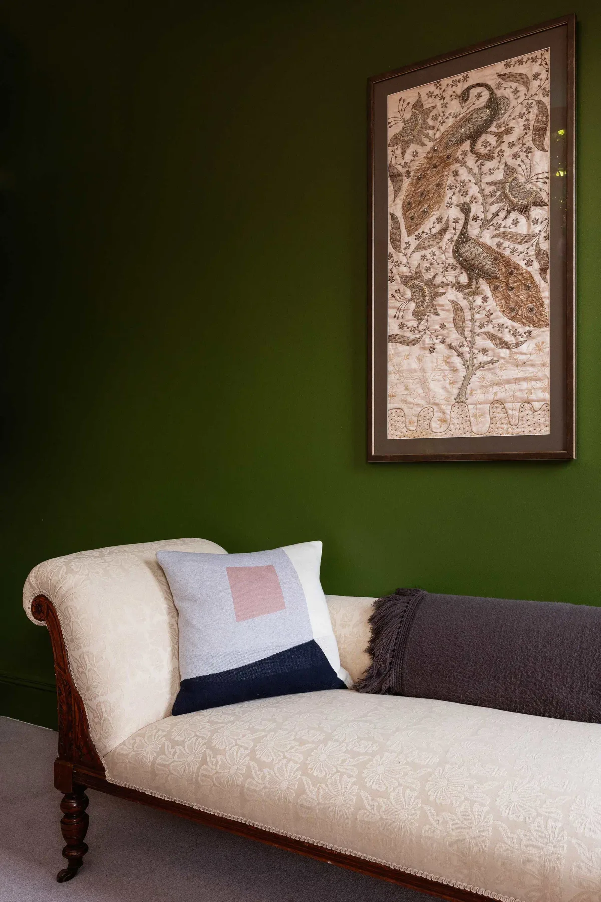 Un asiento tapizado de color crema está junto a una pared verde oscura, bajo un cuadro enmarcado de aves.