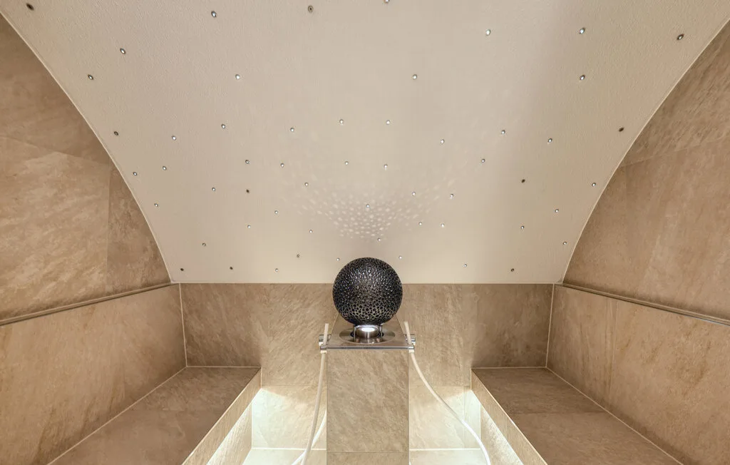 A tiled steam room has a central dark spherical fixture beneath a sloped ceiling.