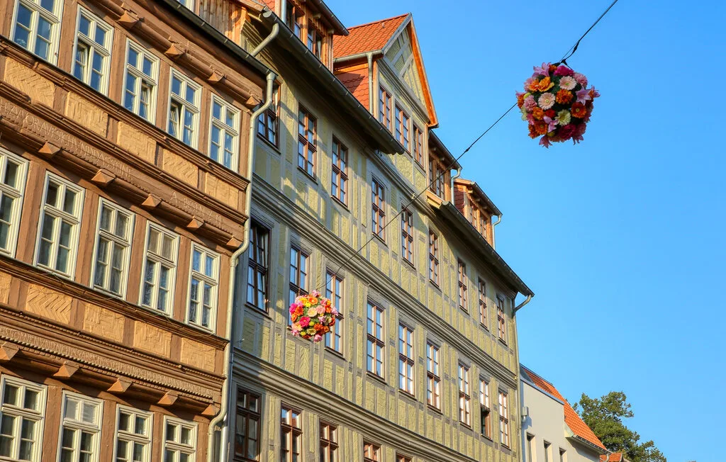 Historic half-timbered buildings line a sunny street beneath hanging flower displays.