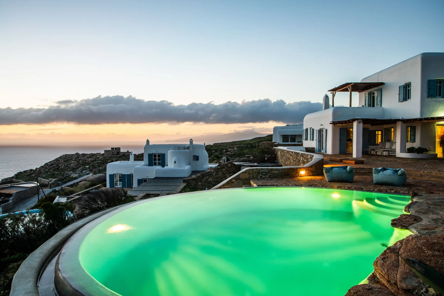 A glowing turquoise pool sits beside white buildings overlooking the coast at sunset.