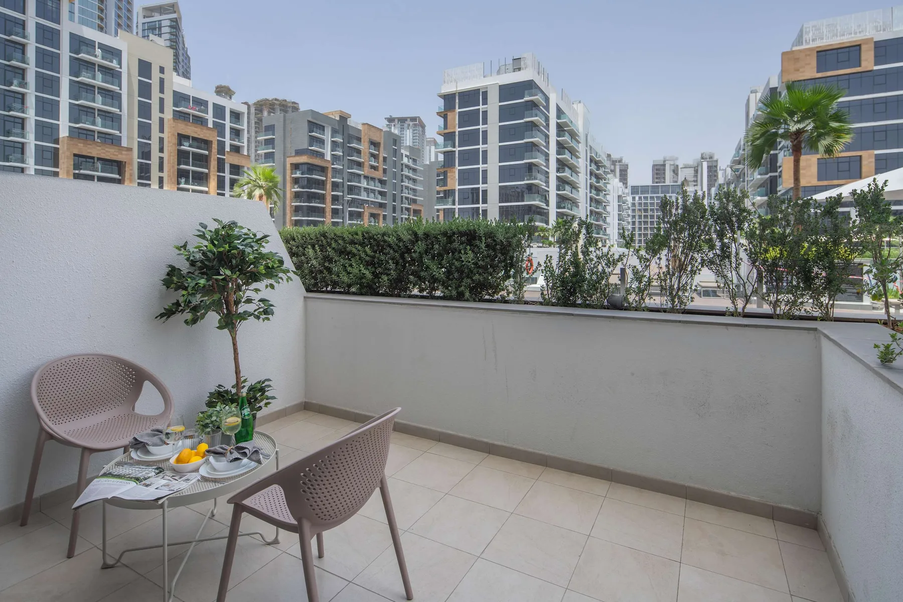 Una terraza exterior con suelo de baldosas, asientos y plantas, frente a edificios cercanos.