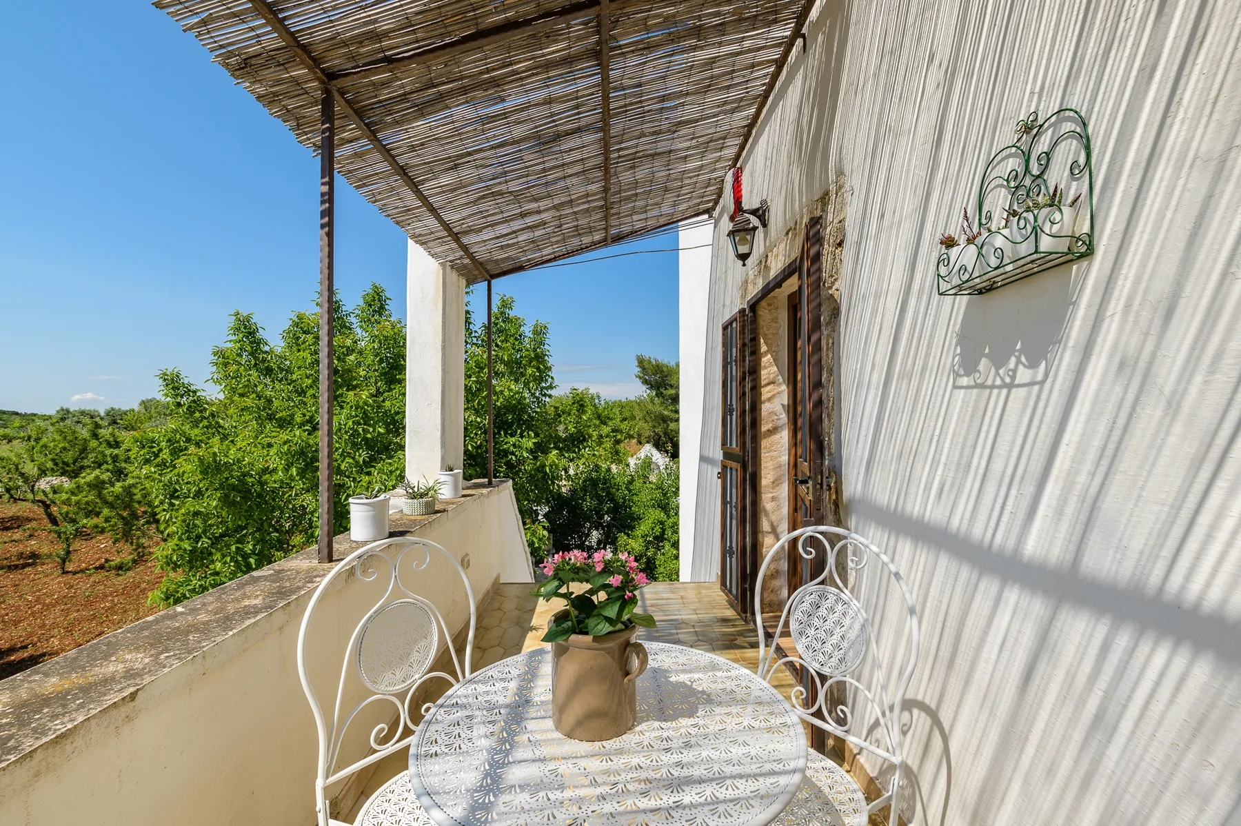 Una pequeña terraza exterior tiene una mesa redonda y asientos junto a una pared blanca.