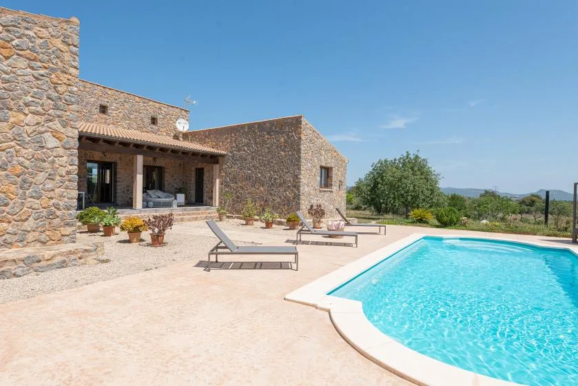 A swimming pool and sun loungers sit beside a stone house on a sunny patio.