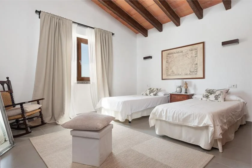 A bedroom with two beds, a curtained window, and exposed wooden ceiling beams.