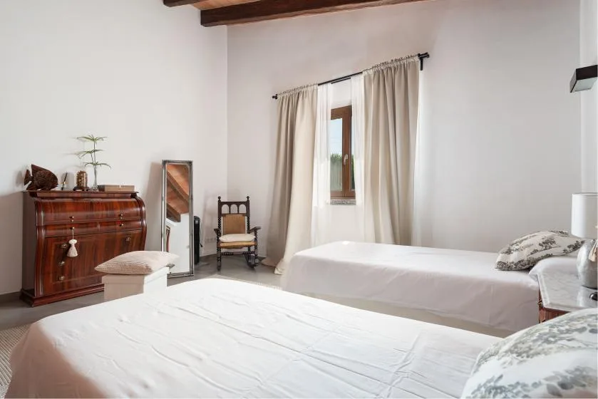 A bright bedroom with two beds, a wooden dresser, and curtains around the window.