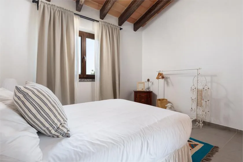 A bedroom with a white bed, beige curtains, and exposed ceiling beams.