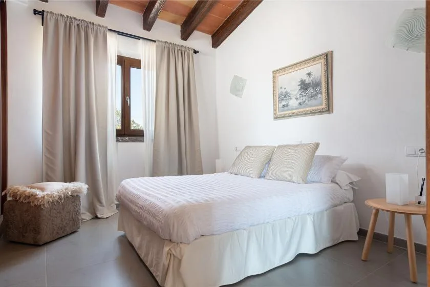A bright bedroom with a neatly made bed, curtained window, and exposed wooden ceiling beams.