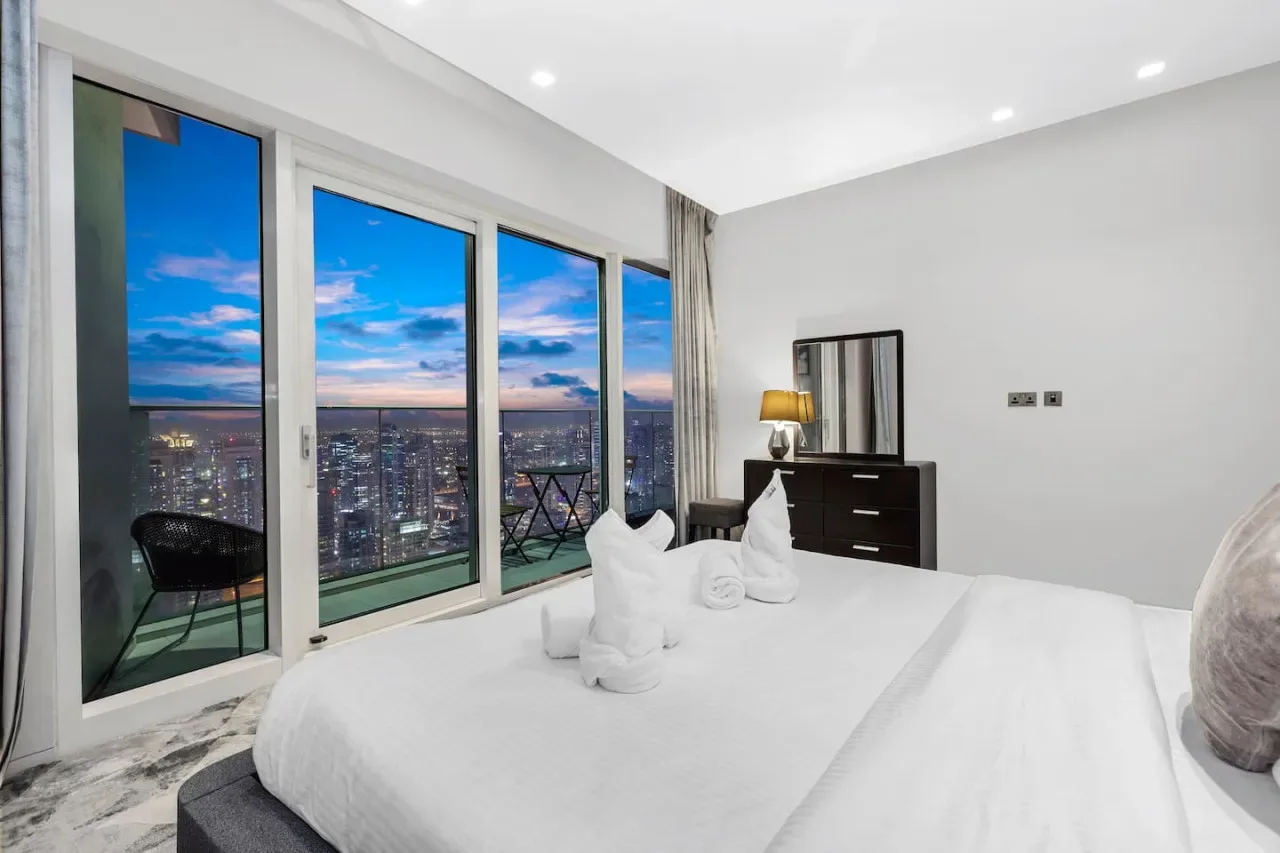 Un dormitorio luminoso tiene una cama grande junto a ventanales con vistas a la ciudad.
