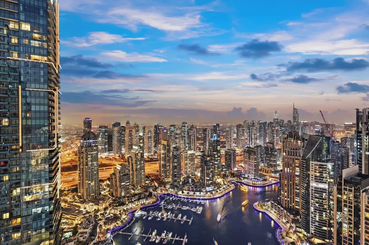Un horizonte urbano al anochecer se alza sobre una marina y un paseo marítimo iluminado.