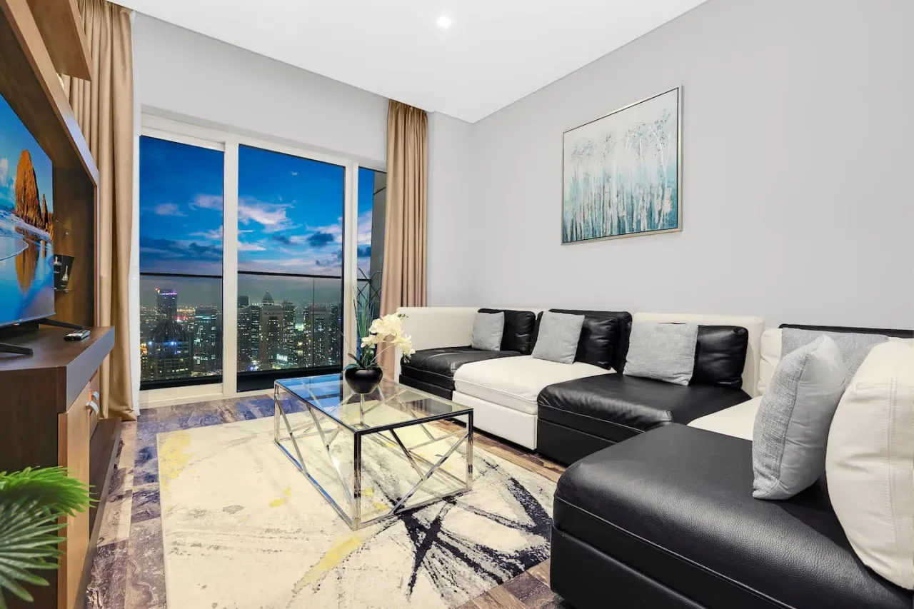Una sala luminosa tiene un sofá seccional, una mesa de centro de vidrio y grandes ventanas con vista a la ciudad.