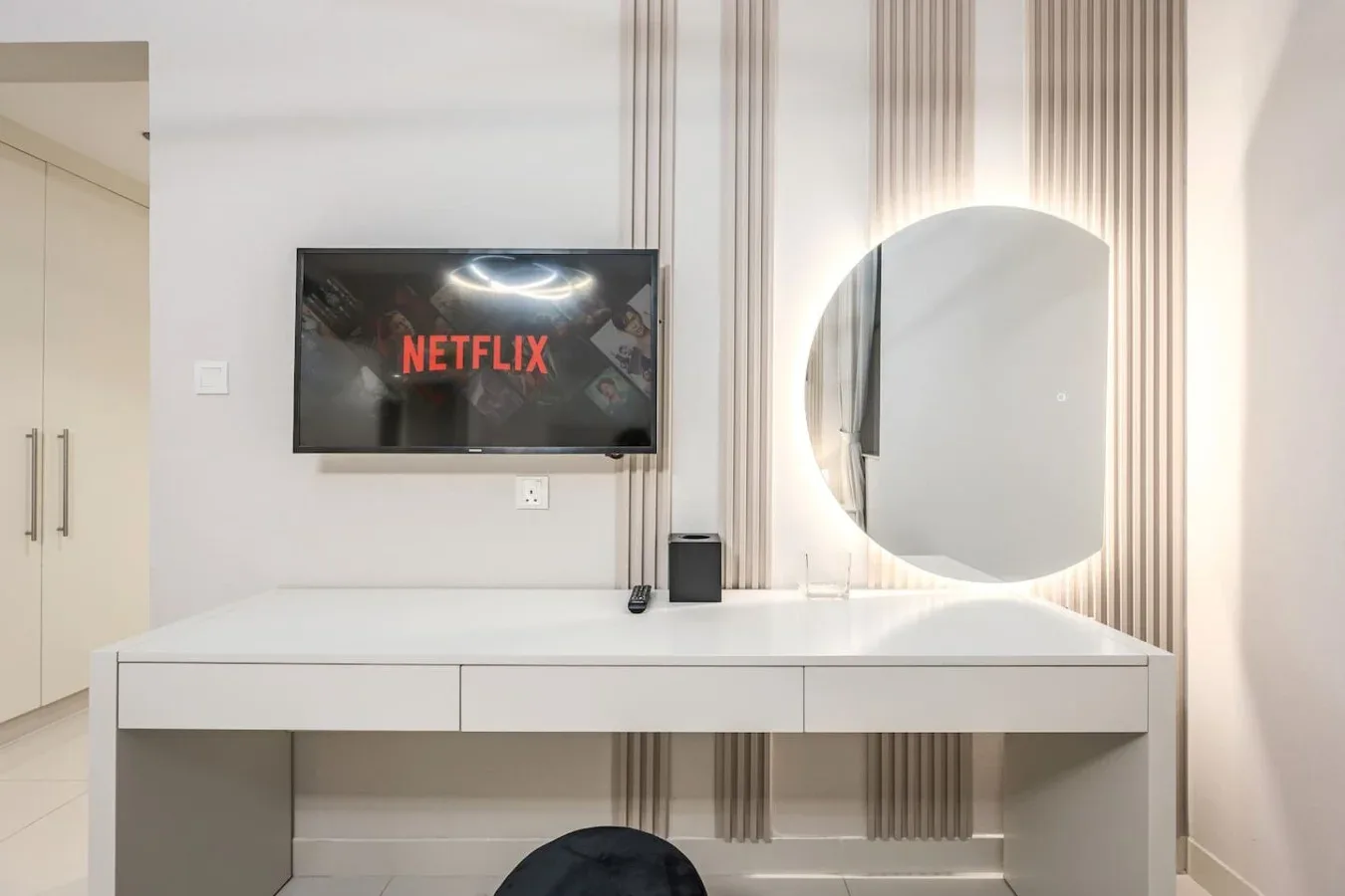 Zona de tocador en un dormitorio con mueble blanco, espejo redondo y televisor en la pared.