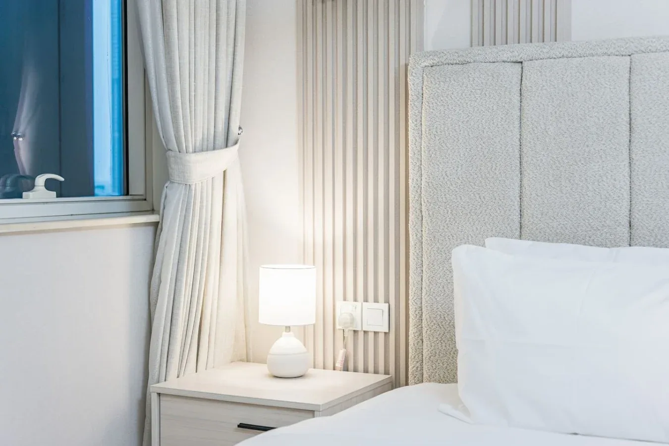 Un dormitorio con una cama junto a una lámpara de mesita encendida y una ventana con cortinas.