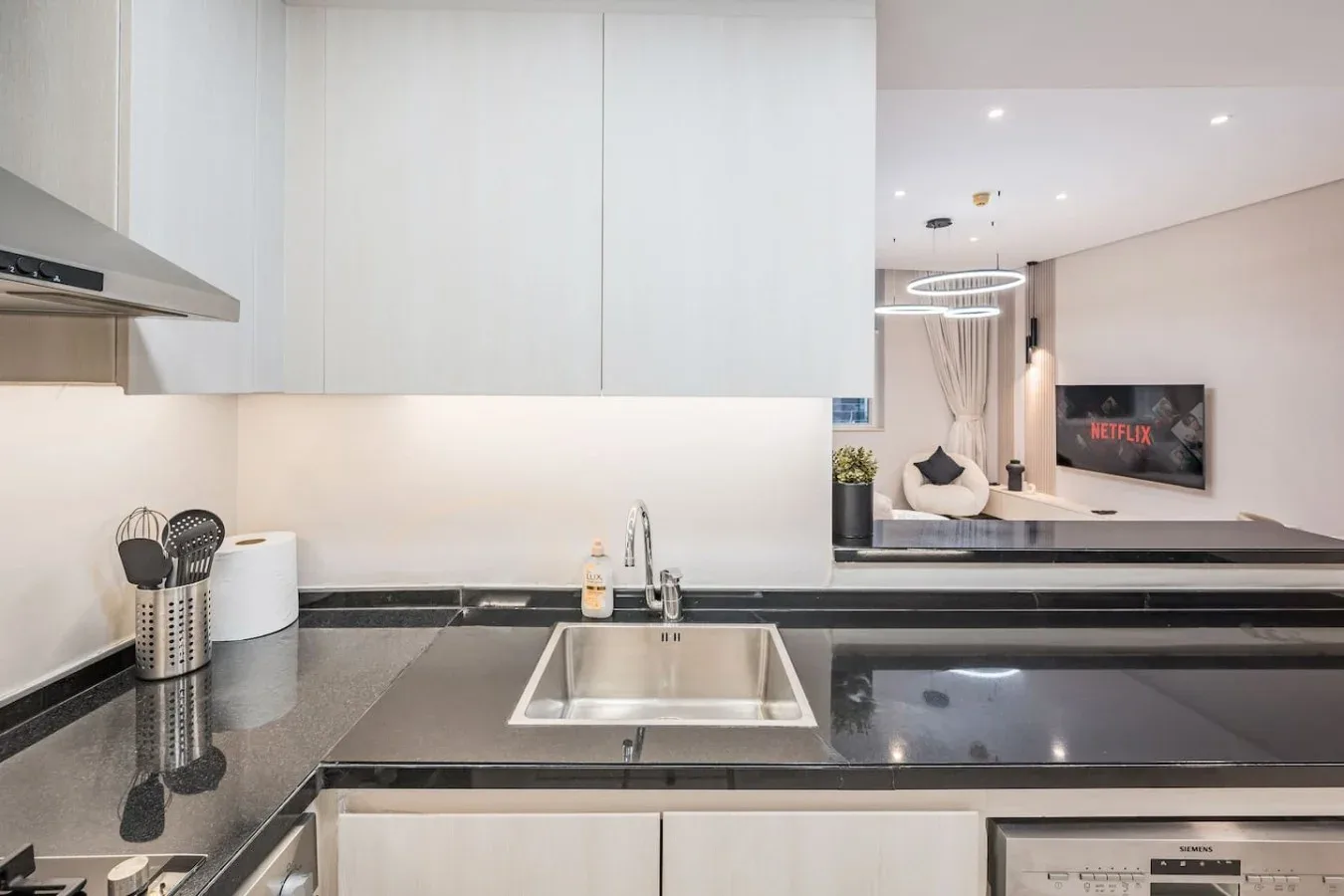 Una cocina moderna con encimeras oscuras y un fregadero de acero inoxidable, abierta a una sala de estar.