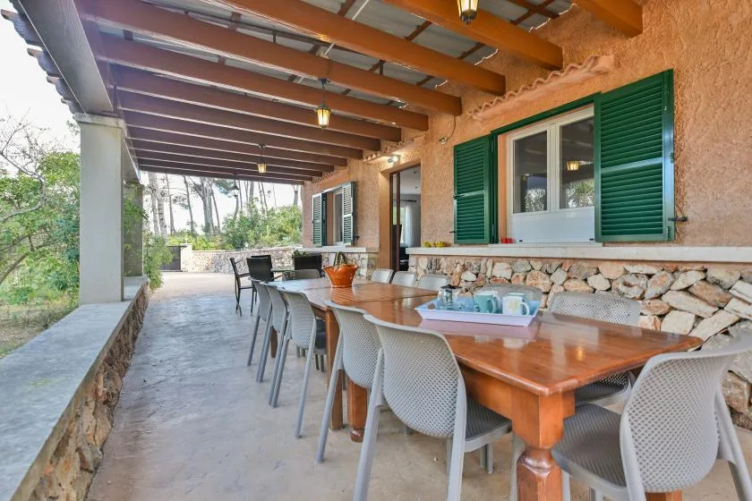 Una zona cubierta para comer al aire libre tiene una mesa larga y varias sillas junto a ventanas con contraventanas verdes.