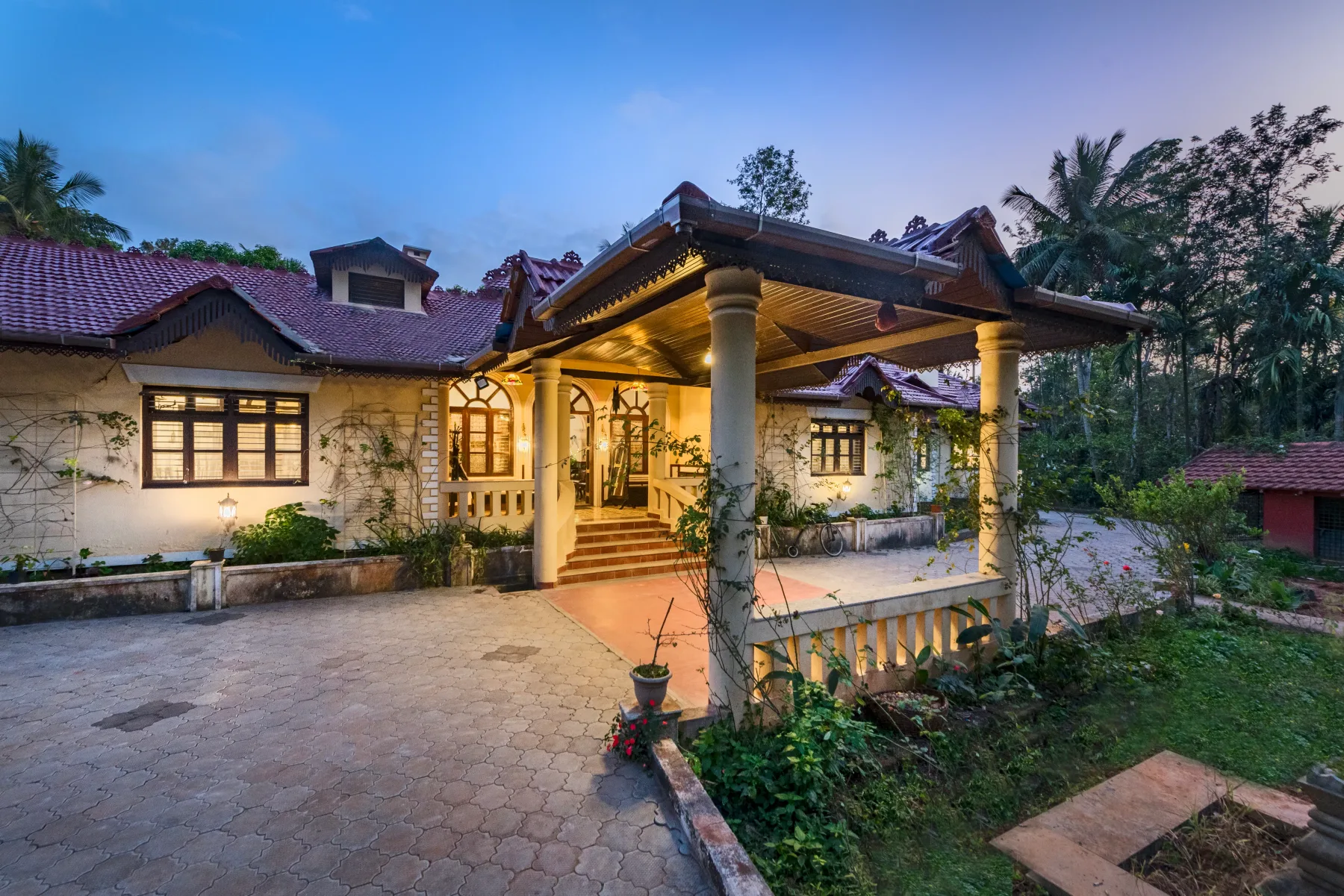 A warmly lit house entrance opens onto a paved courtyard surrounded by greenery.