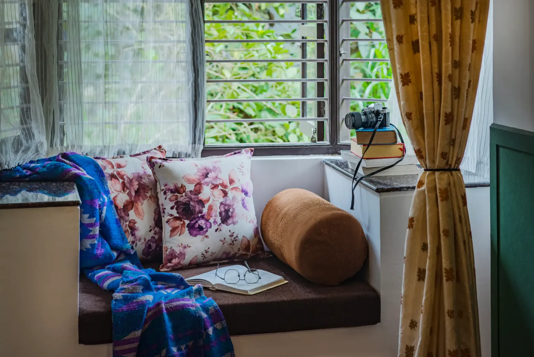 A window-side seating nook has floral cushions and a draped blue blanket.