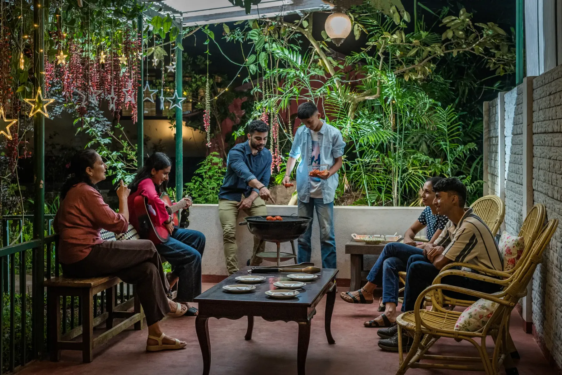 People gather around a barbecue grill in a leafy outdoor seating area.