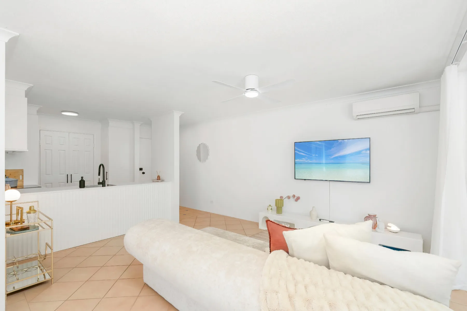 Una sala luminosa con asientos blancos, un televisor y una barra de cocina.