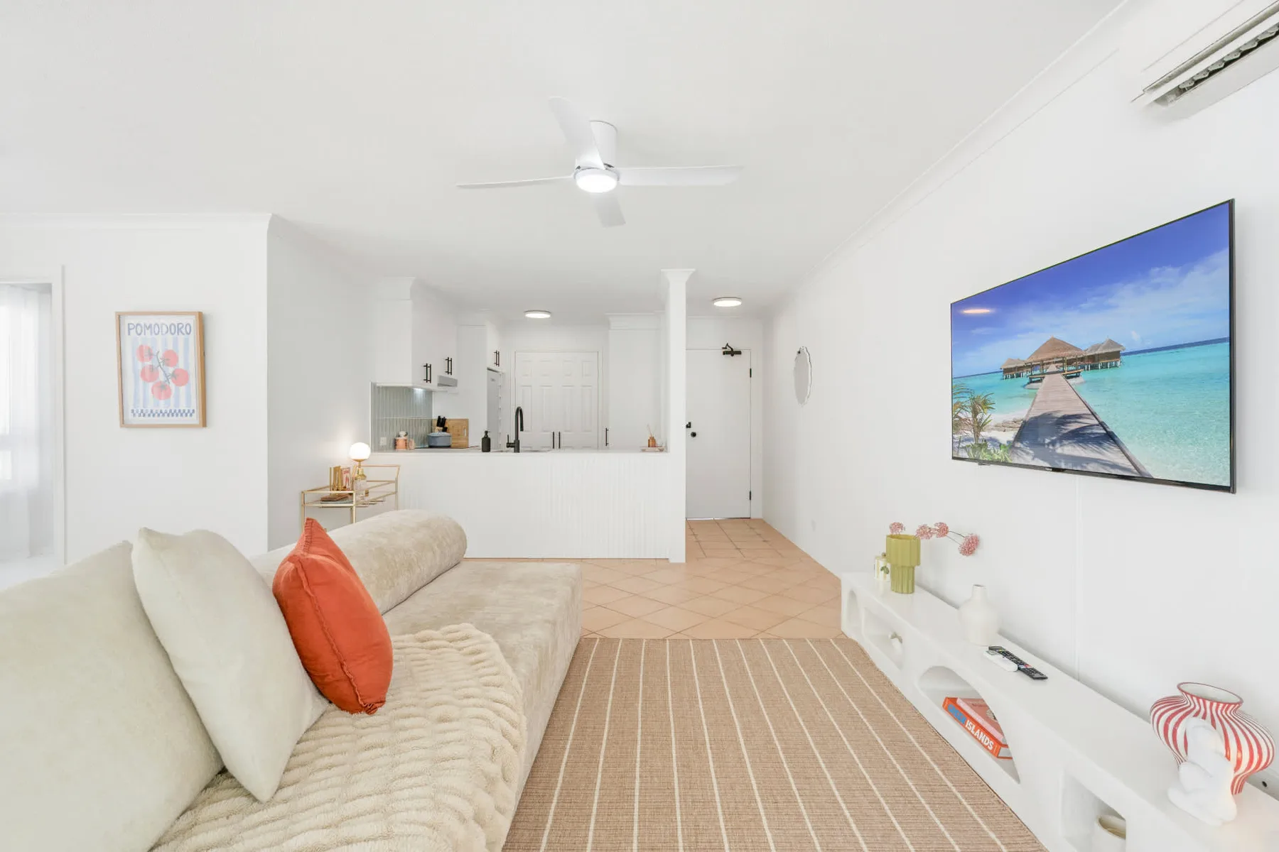 Una sala luminosa con asientos, un mueble blanco para el televisor y una cocina abierta.