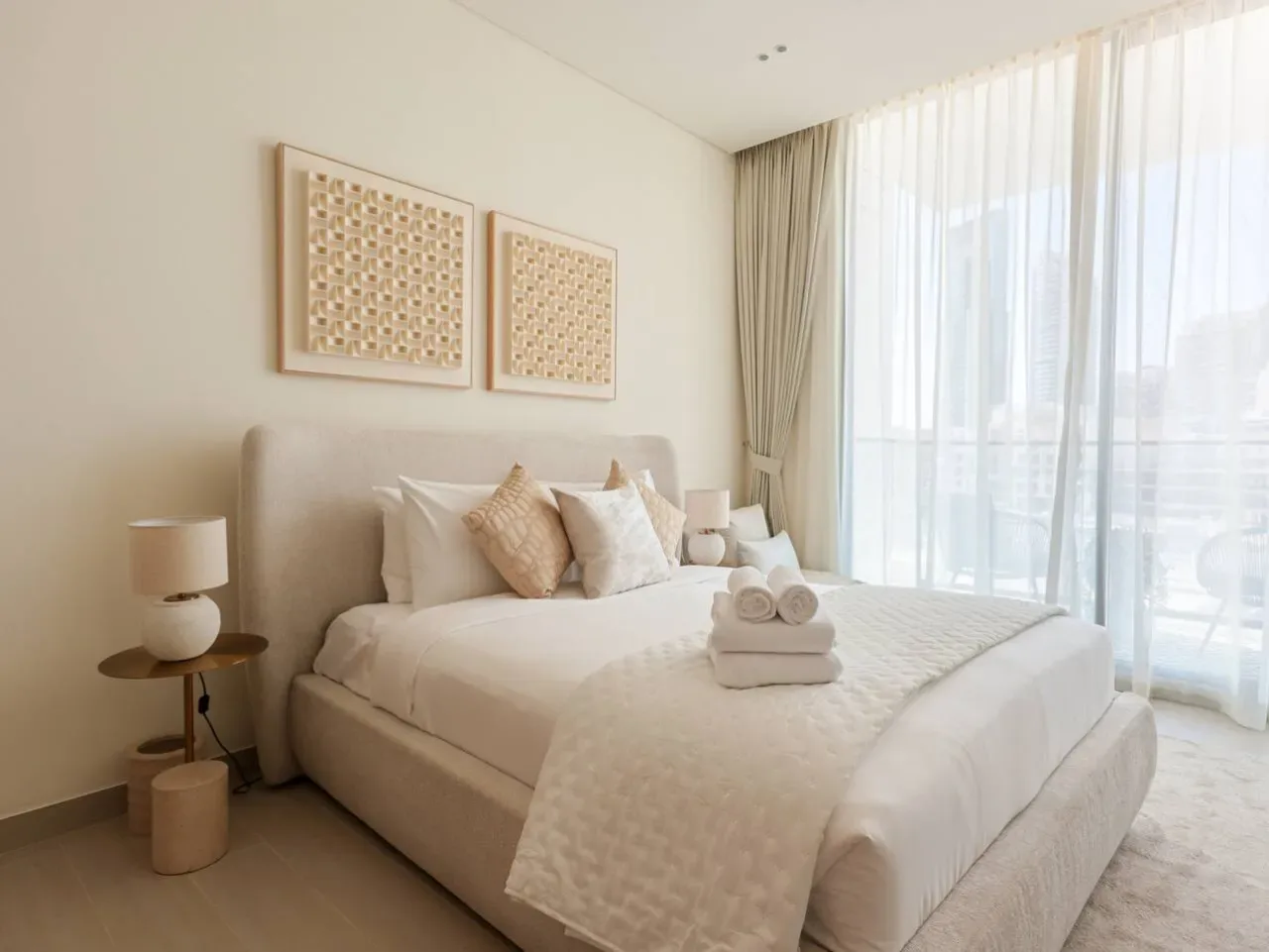 A bright bedroom with a neatly made bed beside large curtained windows.