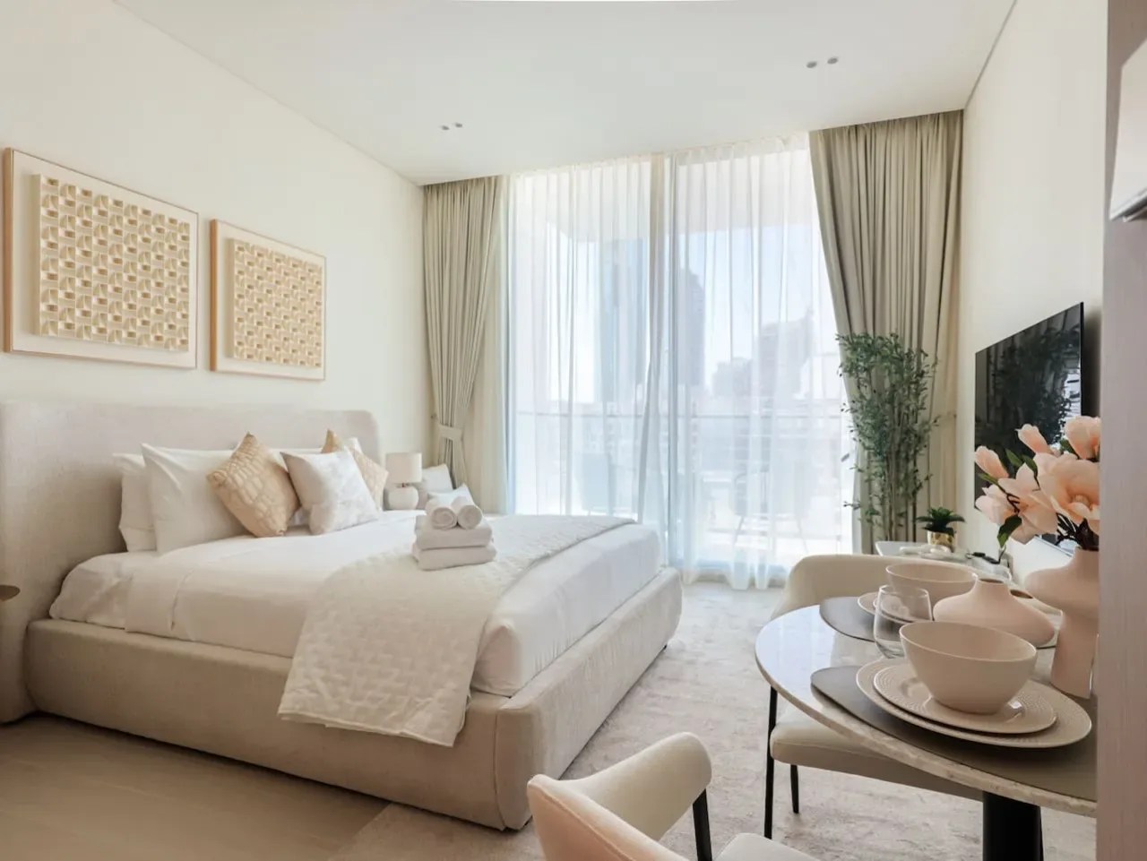 A bright bedroom with a neatly made bed beside large curtained windows.