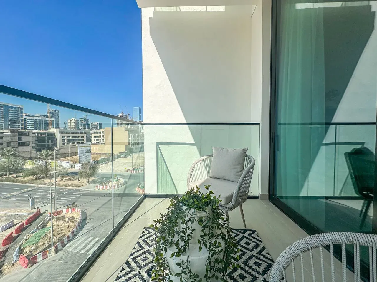 A sunny balcony with glass railings, a small seating area, and a view of nearby buildings.