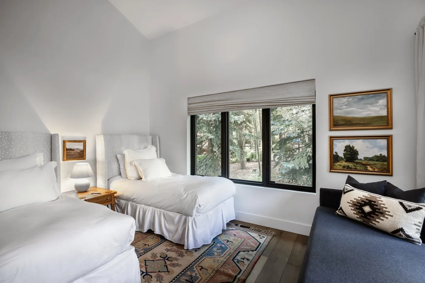 Un dormitorio luminoso con dos camas, una ventana con vista a árboles y una alfombra estampada.