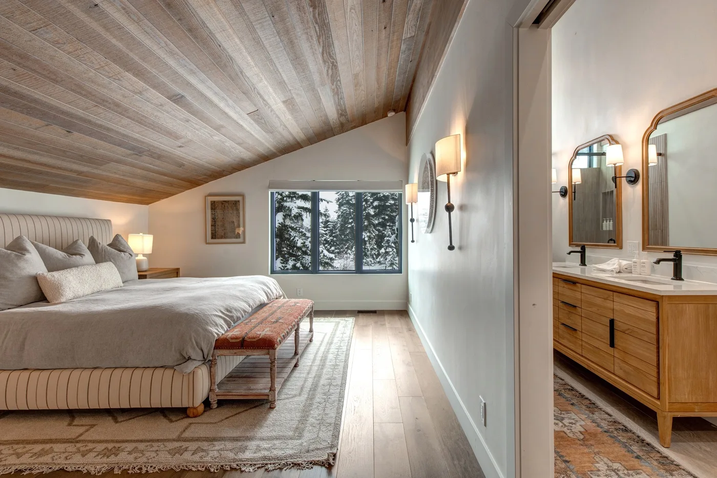 Un dormitorio con una cama grande, techo de madera y un mueble de baño visible por la puerta.