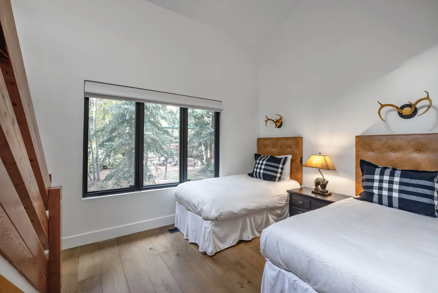 Un dormitorio luminoso con dos camas, una ventana y árboles visibles afuera.