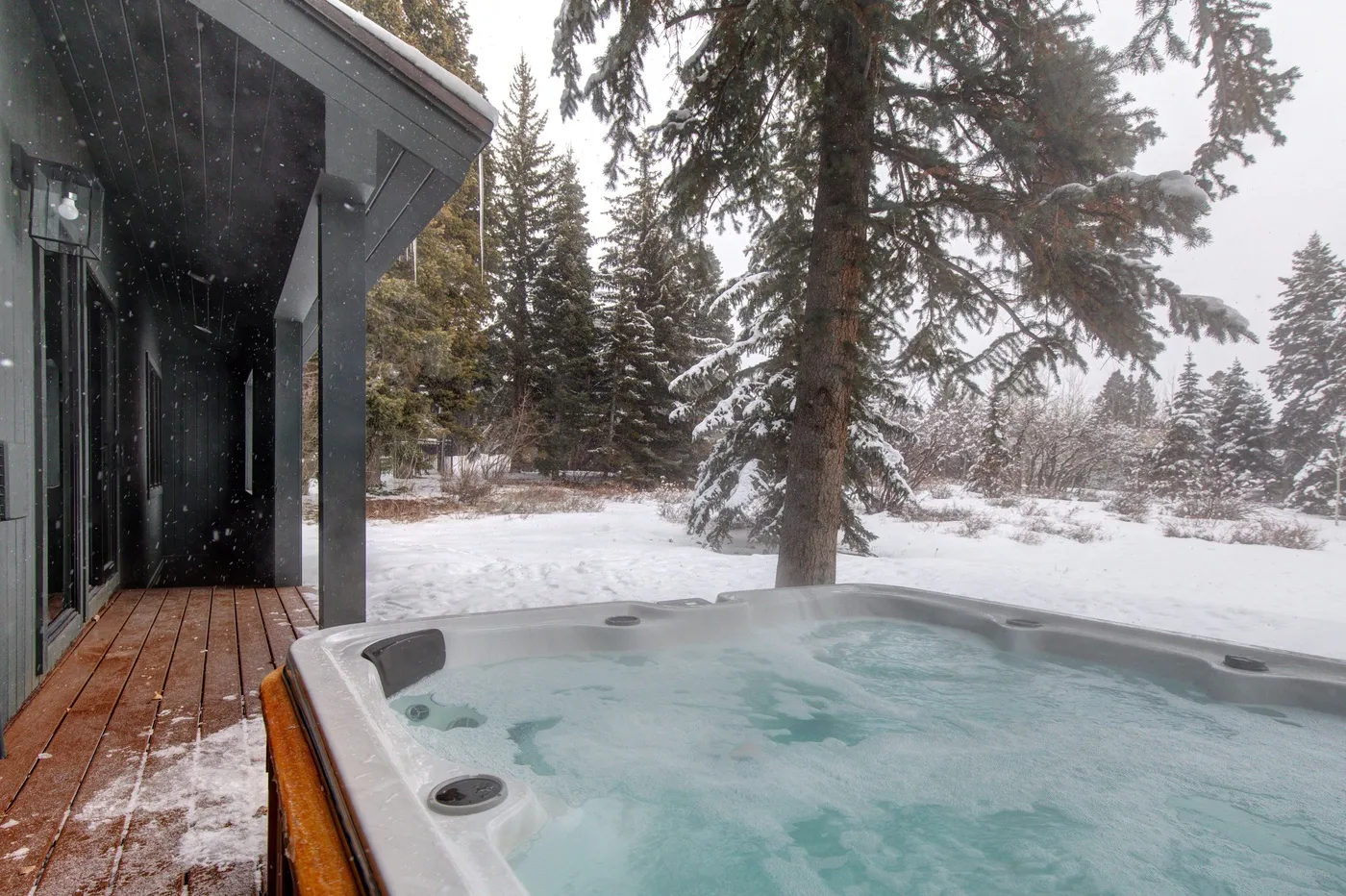 Una bañera de hidromasaje exterior está junto a un edificio moderno en un entorno nevado y arbolado.