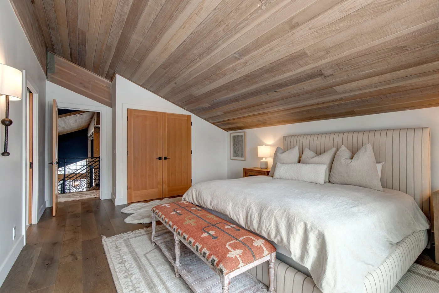 Dormitorio con una cama grande y un banco estampado bajo un techo inclinado de madera.