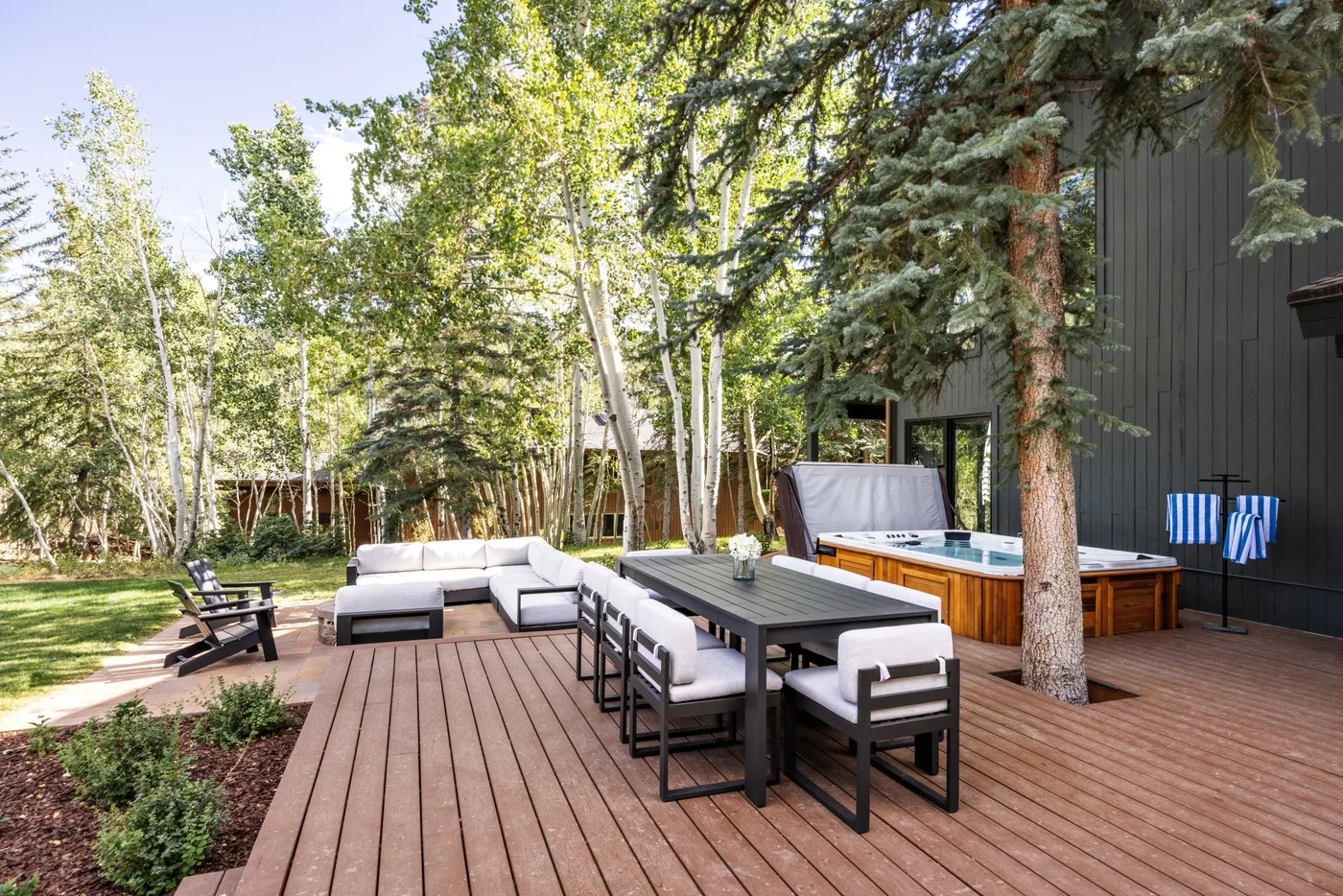 Una terraza de madera con asientos al aire libre y una bañera de hidromasaje está bajo árboles altos.