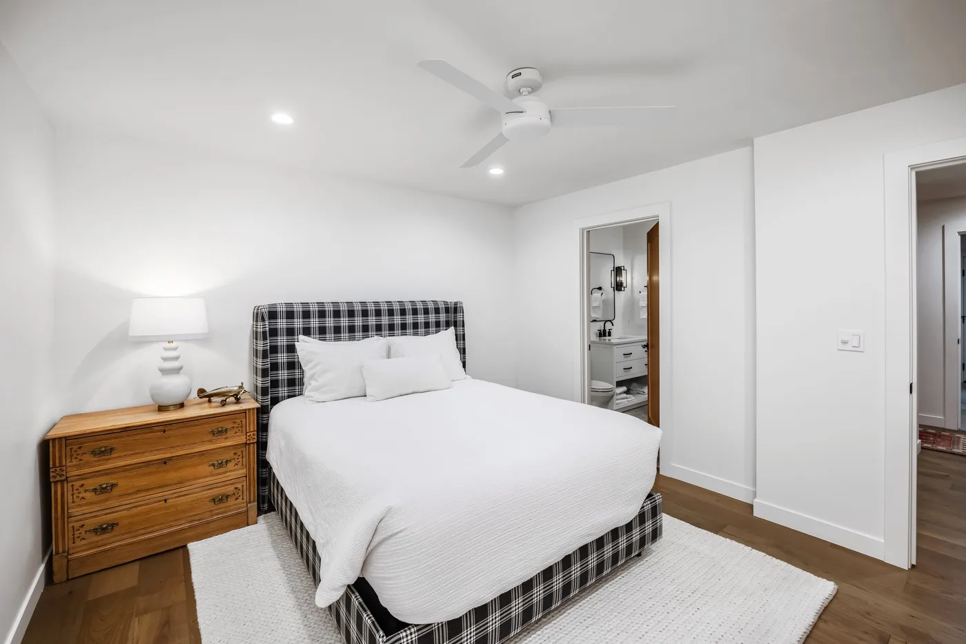 Un dormitorio luminoso con una cama, una cómoda de madera y una alfombra blanca.