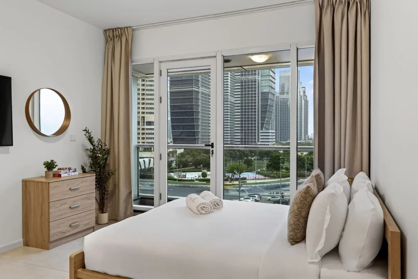 Un dormitorio con una cama bien tendida junto a grandes ventanales con vistas a edificios de la ciudad.