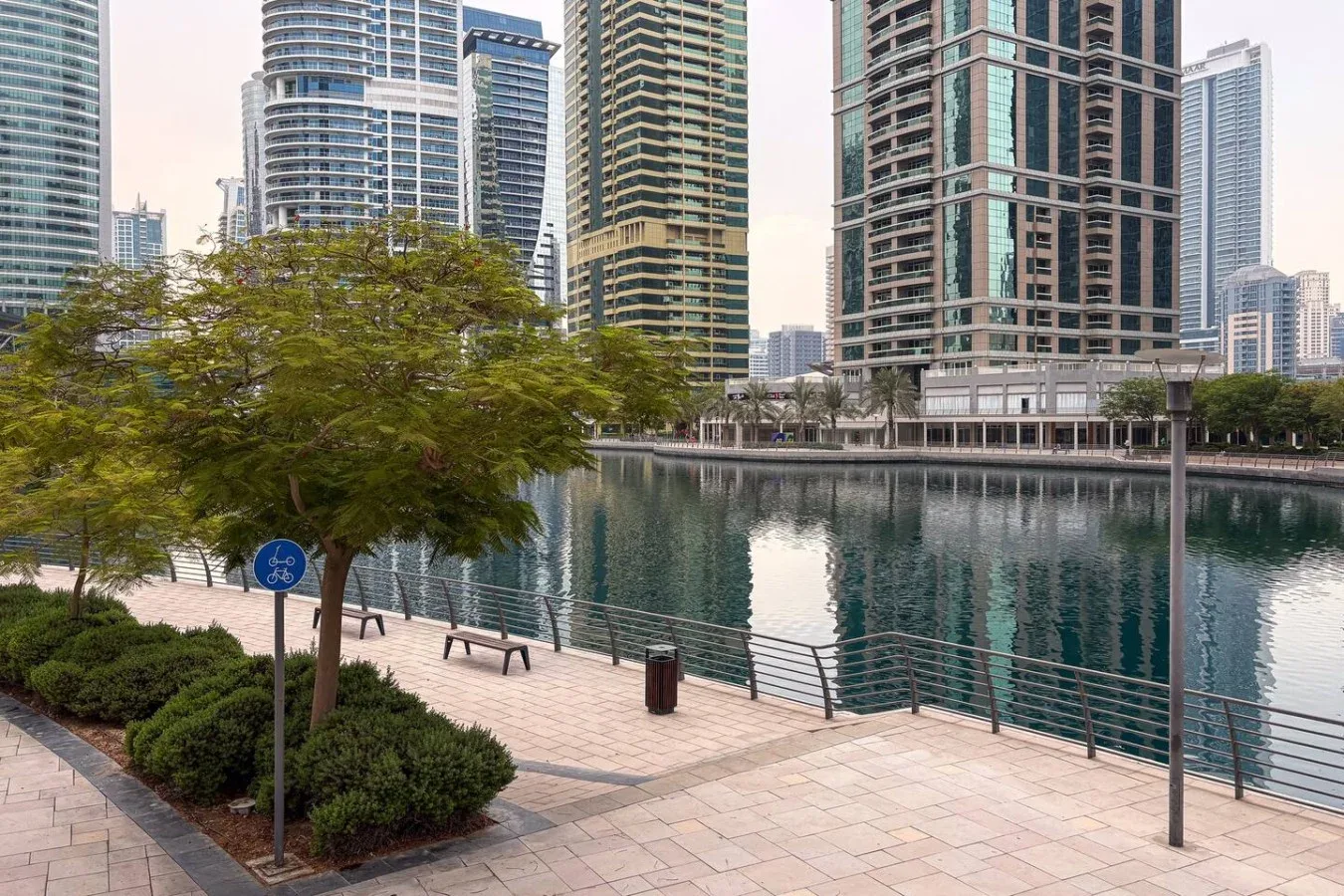Un paseo pavimentado junto al agua bordea un canal, con árboles y edificios altos cerca.