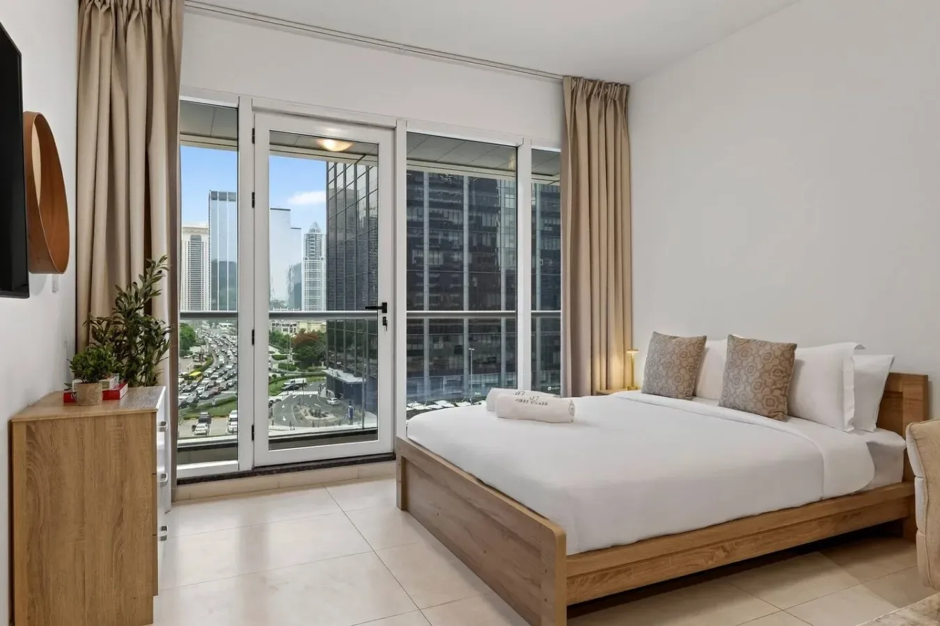 Un dormitorio luminoso con una cama junto a grandes ventanas con vistas a edificios cercanos.