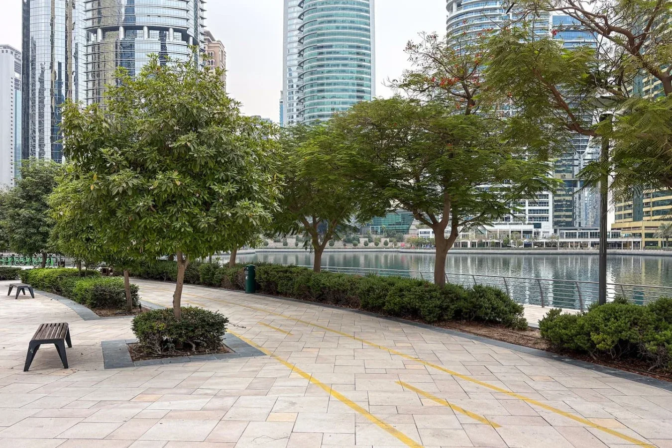 Un paseo pavimentado junto al agua pasa bajo los árboles, con edificios altos al otro lado.