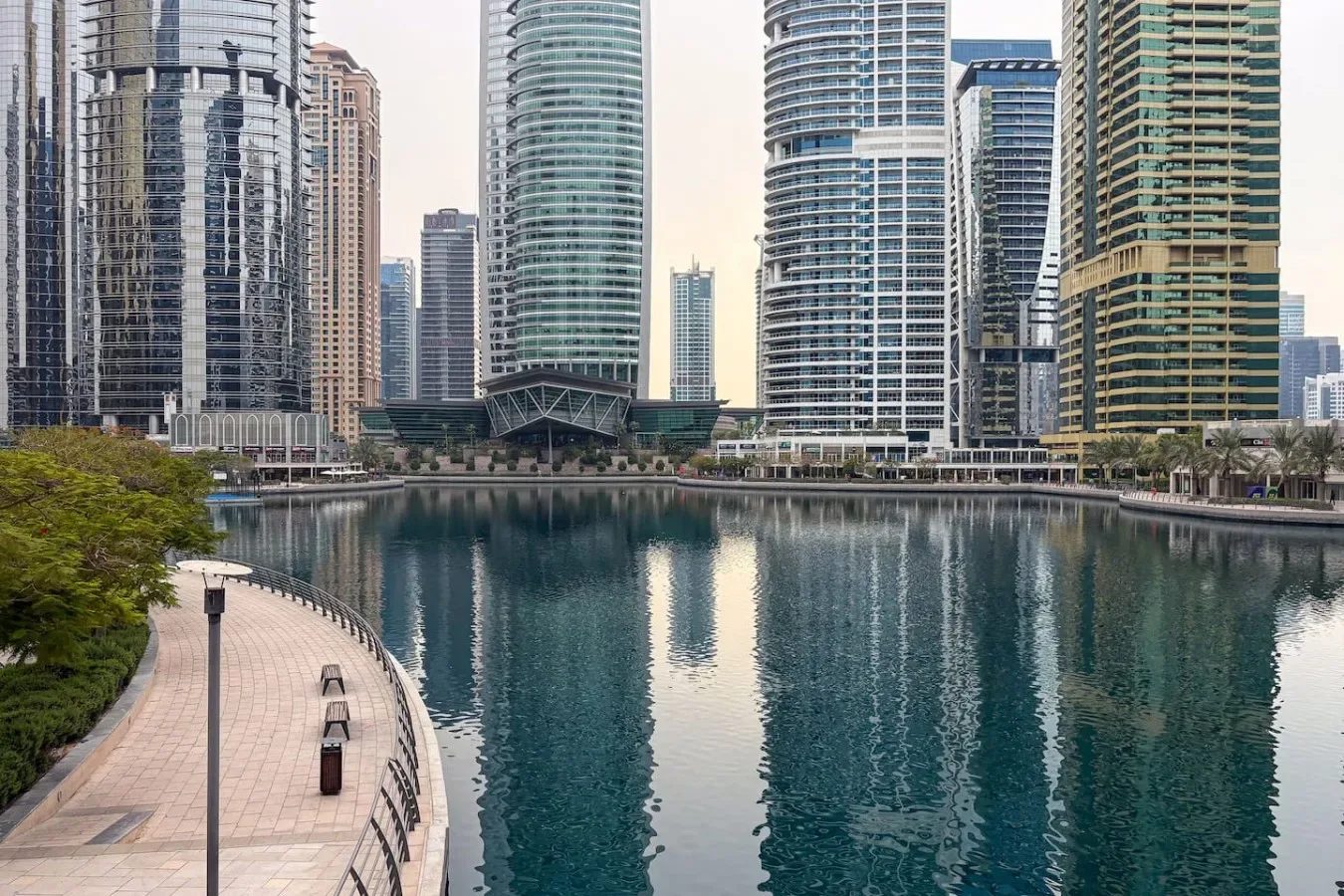 Un paseo junto al agua bordea un canal, con modernos edificios altos al fondo.