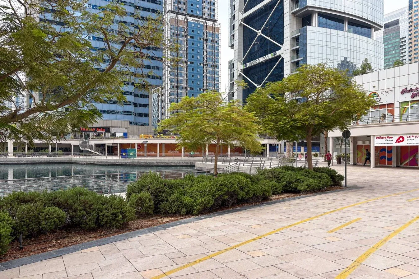 Una plaza al aire libre junto a un espejo de agua, con árboles, pavimento y edificios alrededor.
