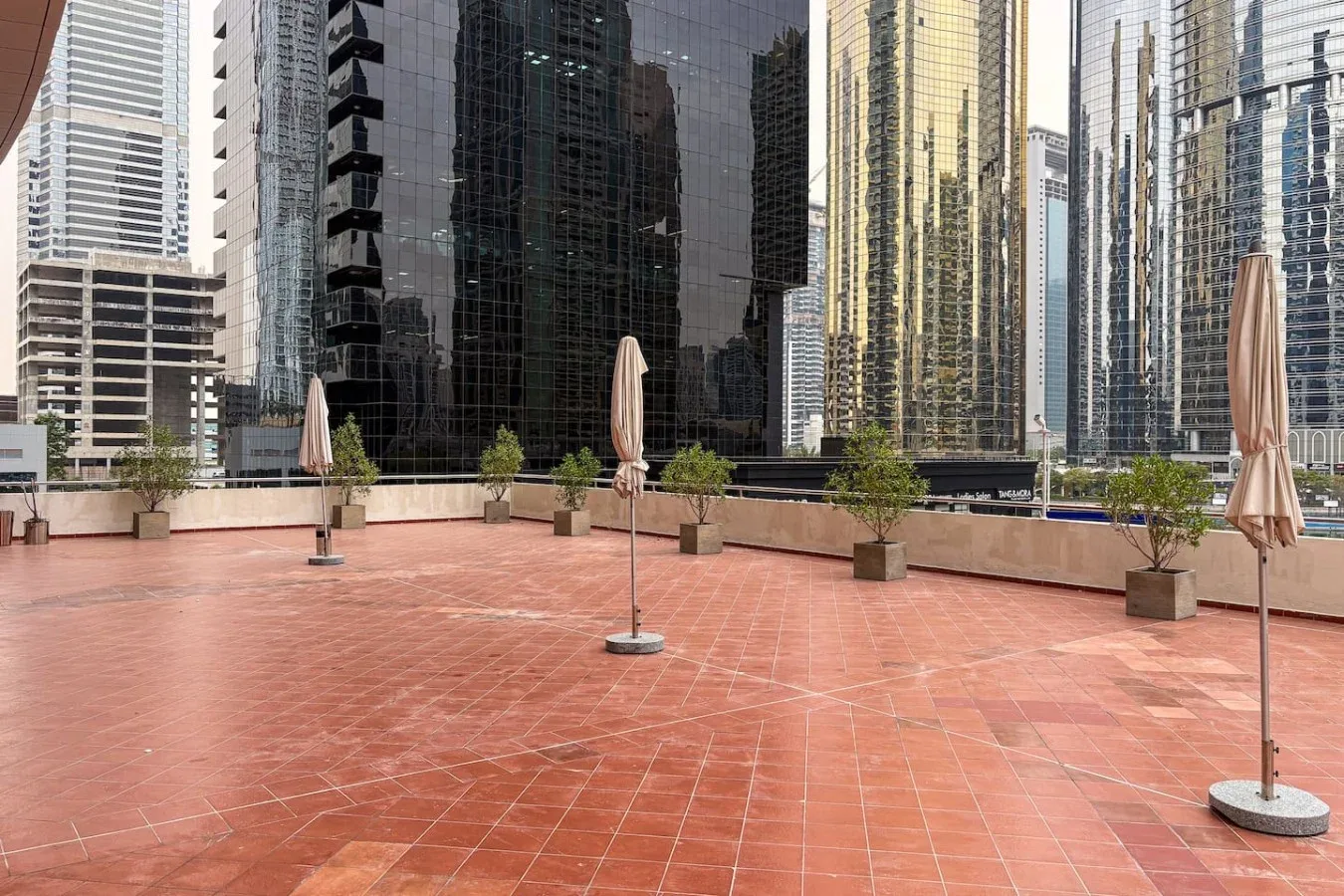 Una terraza en la azotea con baldosas de terracota, plantas en macetas y sombrillas cerradas da a edificios altos.