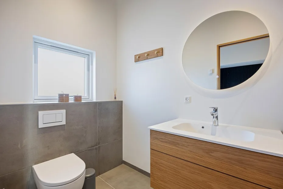 Un baño moderno con inodoro suspendido, mueble de madera y espejo redondo.
