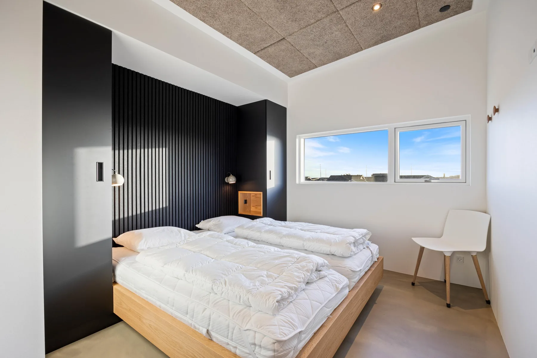 Dormitorio con una cama baja, ropa de cama blanca y dos ventanas.