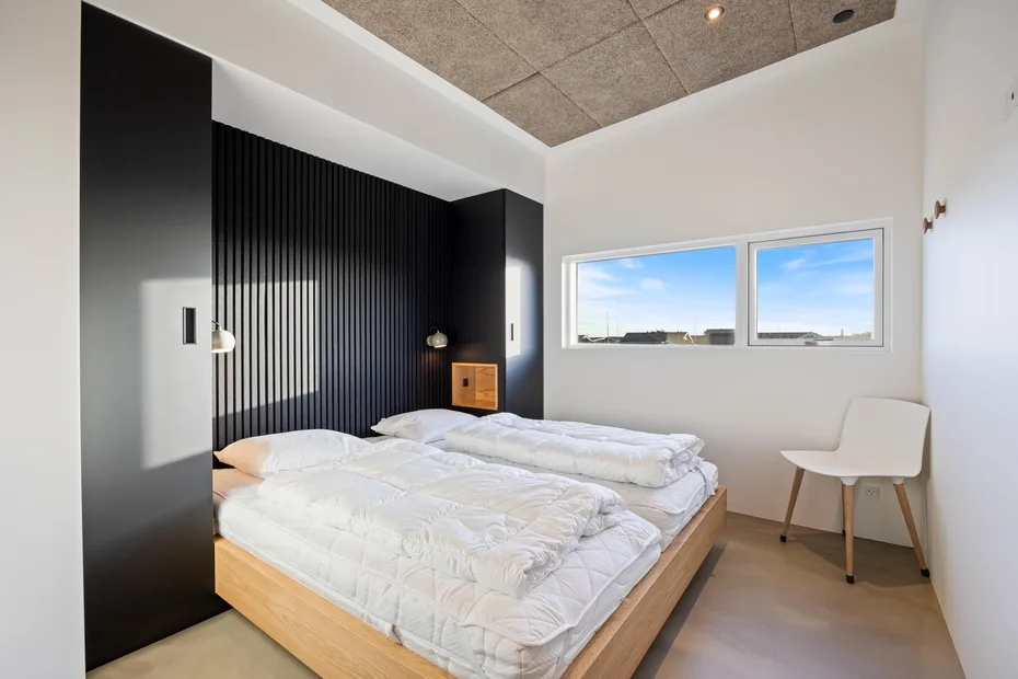 Un dormitorio con una cama baja, ropa de cama blanca y dos ventanas.