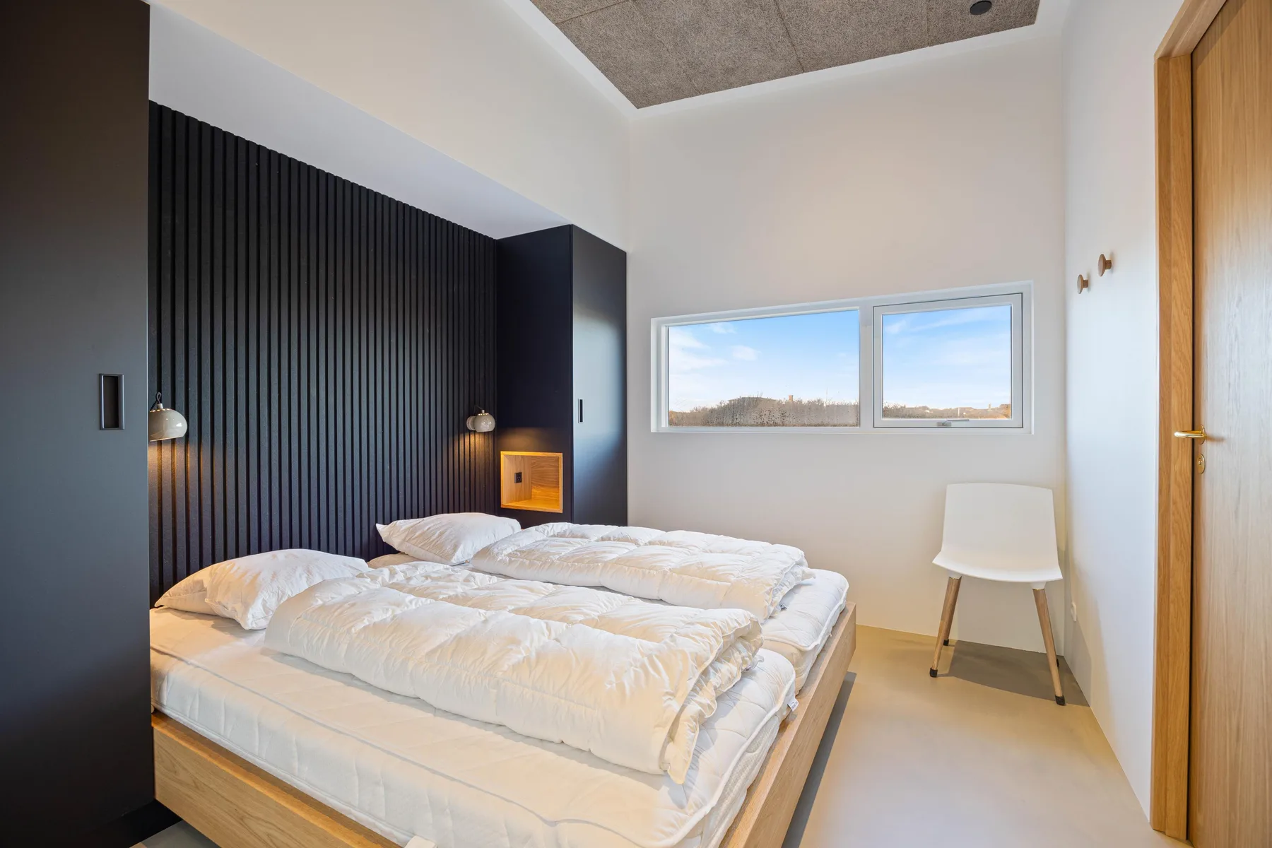 Un dormitorio con una cama, una pared oscura con paneles y una ventana amplia.