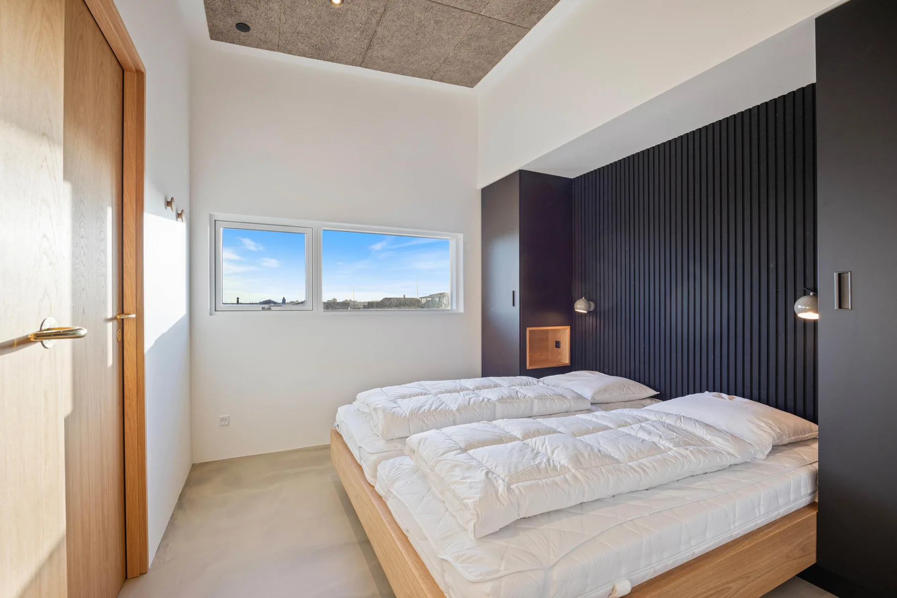 Un dormitorio luminoso con una cama baja, ropa de cama blanca y ventanas amplias.