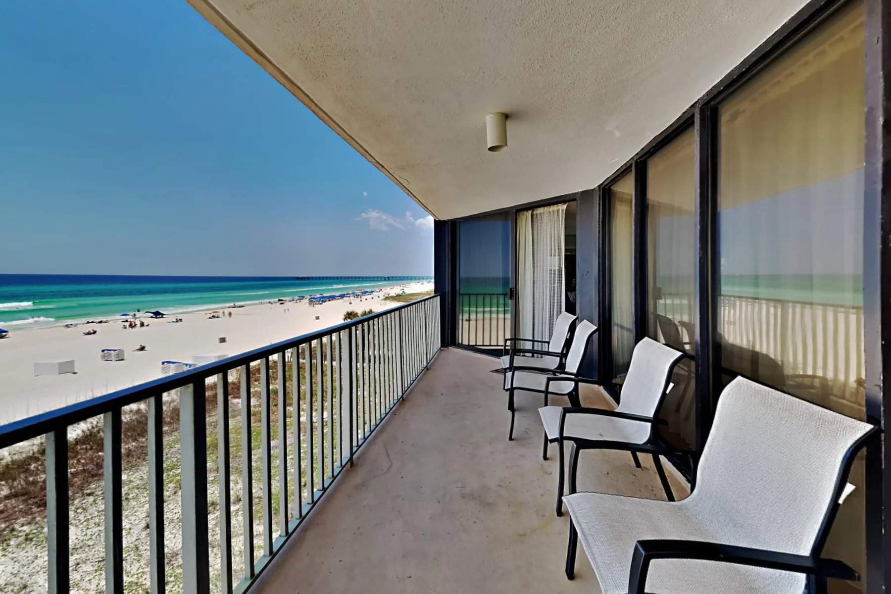 A covered balcony overlooks a sandy beach and ocean, with seating along the glass doors.