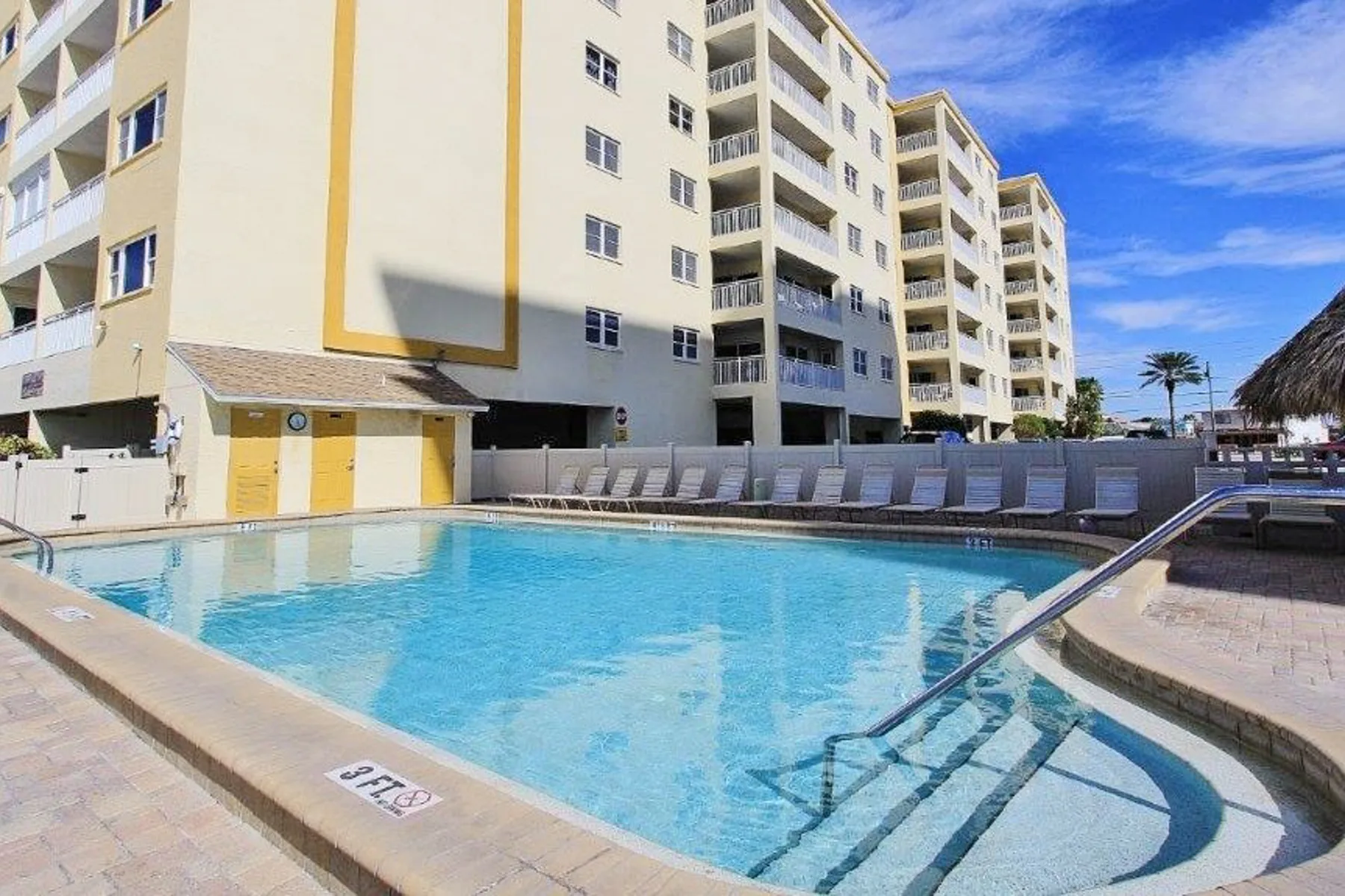 An outdoor swimming pool sits beside a multi-story building under a blue sky.